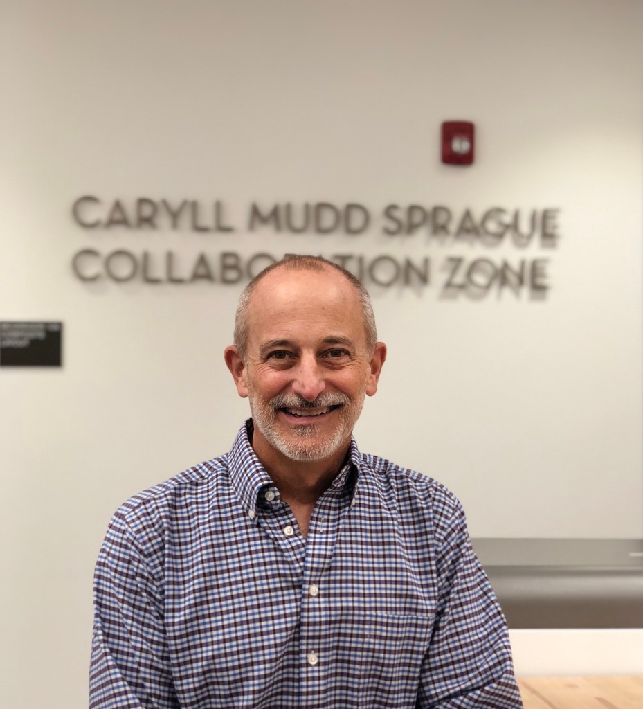 An image of Professor Groves, a white man with short hair and a white beard wearing a checkered shirt. He is sitting in front of a sign that says "Caryll Mudd Sprague Collaboration Zone".